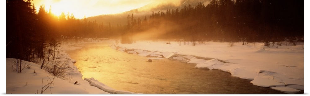 Panoramic image of a river running through a frozen landscape with fog coming off it and warm sunlight breaking through th...