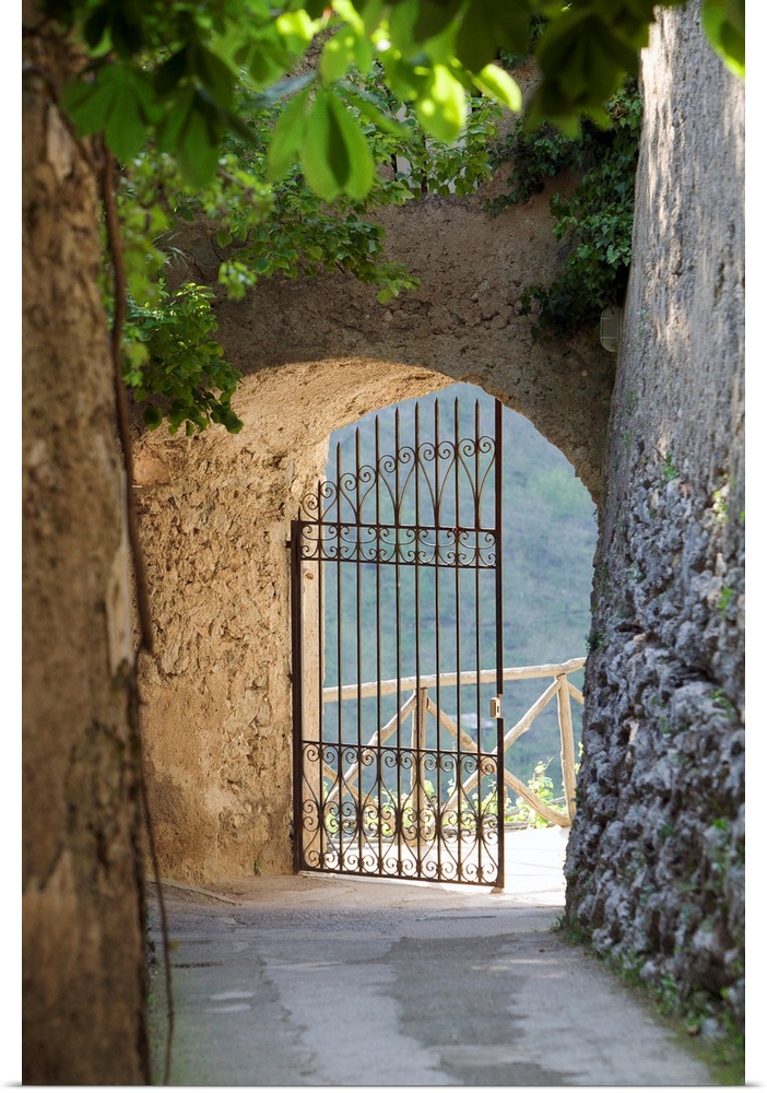 A vertical photograph of a walkway lined with old masonry a green vines to an open iron wrought gate.