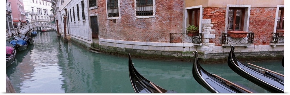 The front of gondola boats are pictured in the water with buildings to either side of the canal where more boats are shown.