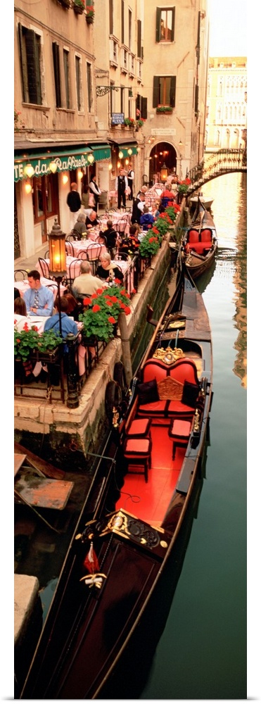 Vertical panoramic photograph boats on canal waterway lined with street cafes and buildings.