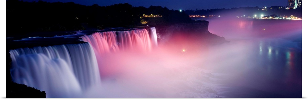 Panoramic photograph of wide stretching waterfall at night with fog rising from the bottom.  There are mountains and city ...