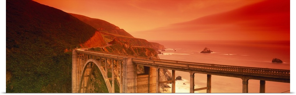 Oversized, horizontal photograph of the Bixby Bridge, next to the cliffs of Big Sur, California, a fiery sunset casts a wa...
