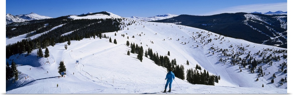 A panoramic photograph of a skier departing down a snow covered slope at the top of a mountain.