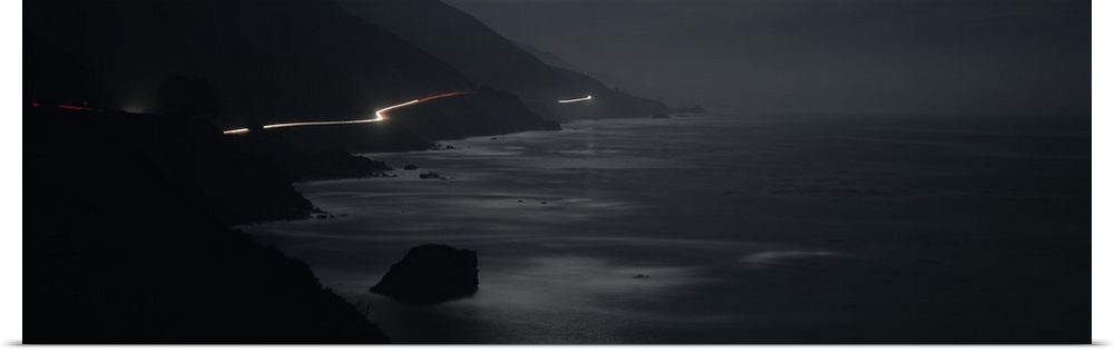 Panoramic photograph of road near coastline lit up with moving headlights at night.