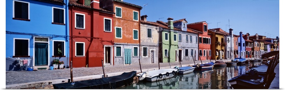 Panoramic photograph of colorful houses lined up next to each other on a lagoon with boats tied up in the water.