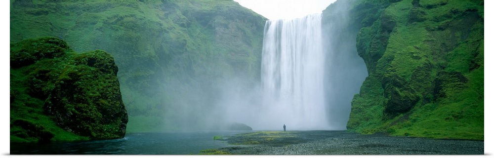 Panoramic photo of a wide waterfall spilling over a cliff into the water below with a man standing on the land in front.