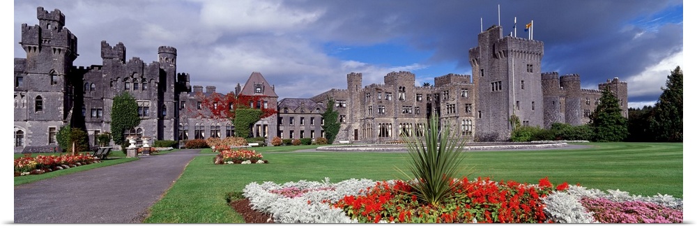 Panoramic photograph taken of an immense castle that has lush green grass in the front yard filled with planted flowers.