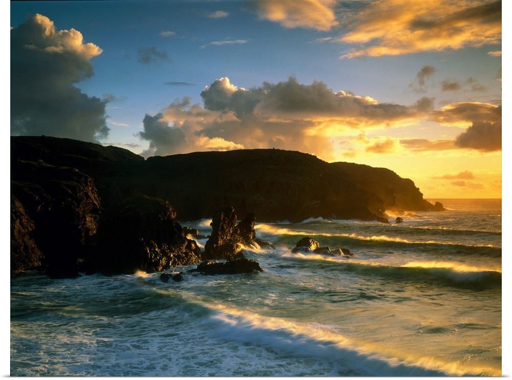 Photograph of rock cliffs in ocean with waves rolling in under a cloudy sky at sunrise.