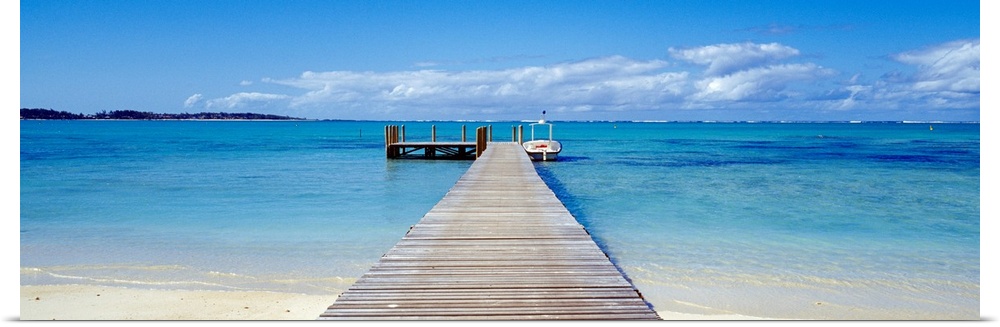 Panoramic photograph of pier stretching into the sea off of a sandy beach.  There is a small wooden deck at the end of dock.