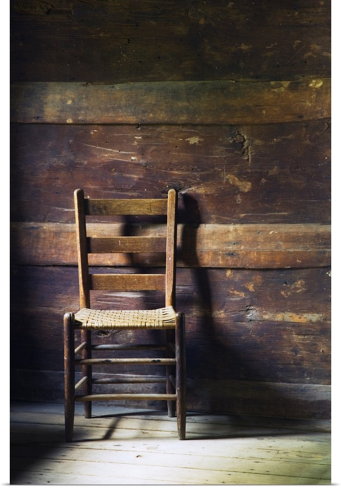 Portrait photo of an empty ladderback wooden chair sitting in a beat up wood slat wall room with sanded down floors.