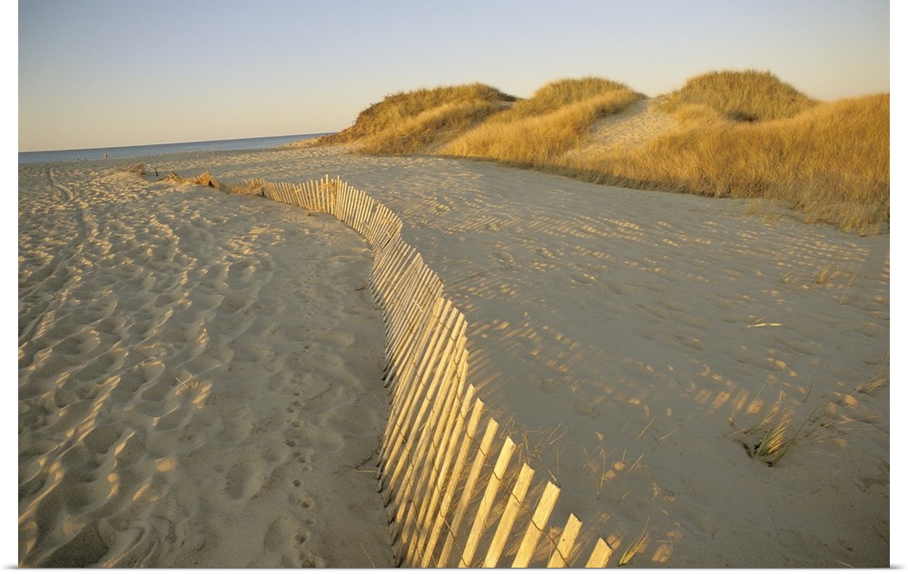 Large photograph taken of a sandy beach that has a fence running through the middle of the picture.  In the background the...