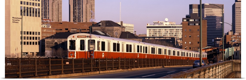 An above ground subway train is photographed in panoramic view with the Boston skyline shown in the background.
