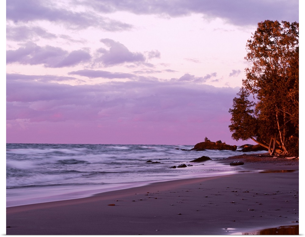 Picture of waves in Lake Superior crashing on to the beach as the sun sets and lights up the sky in purple tones.
