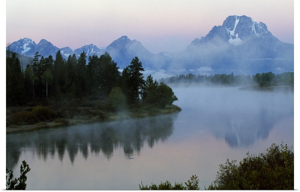 A large photograph showing forest and foilage surrounding water with mountains in the background and mist floating above.