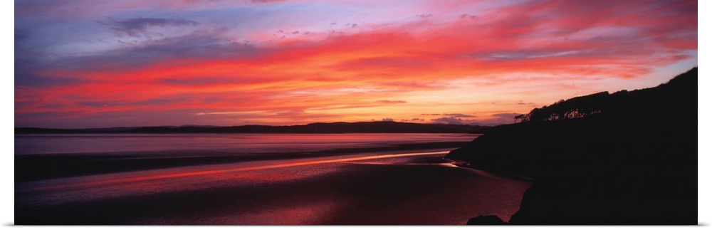 A large panoramic photograph taken of a sunset over a bay in the UK. A hill top is silhouetted to the very right of the pr...