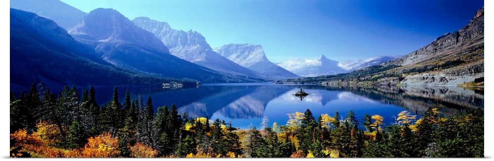 A panoramic photograph of mountains reflecting in a still lake surrounded by trees in this landscape of the wilderness.