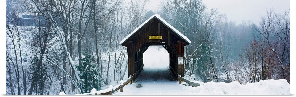 A panoramic photograph of a one lane road through a covered bridge in a winter snowscape.