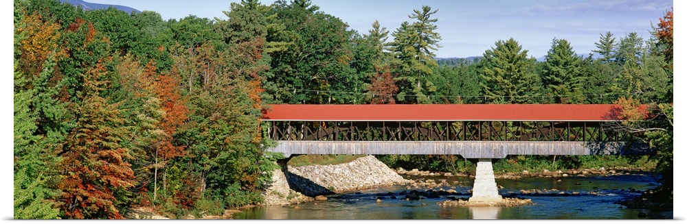 Panoramic image of a red roofed covered bridge spanning a calm tree lined river.