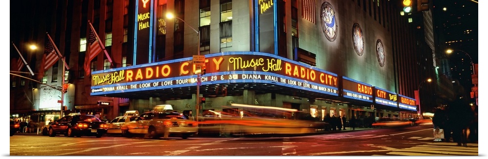 Panoramic view of Radio City Music Hall from the street corner at night, lit up by neon lights and with taxis passing by.