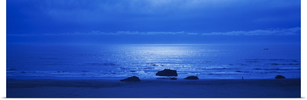 Panoramic photograph of a monotone seascape with the moonlight reflecting off the waves, three solitary rocks sit on the s...