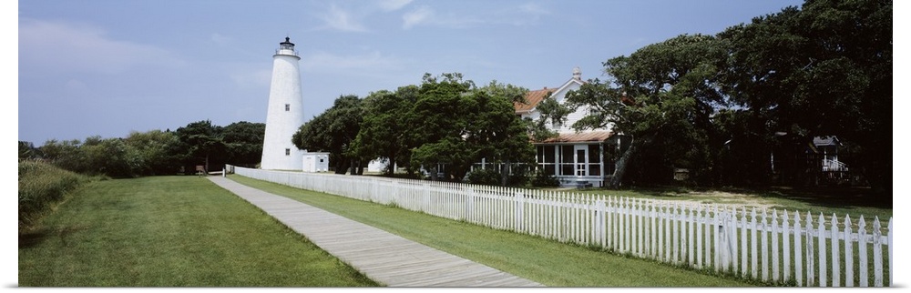 Horizontal photograph on a big canvas of a thin boardwalk and fence leading to a lighthouse surrounded by trees, a house i...