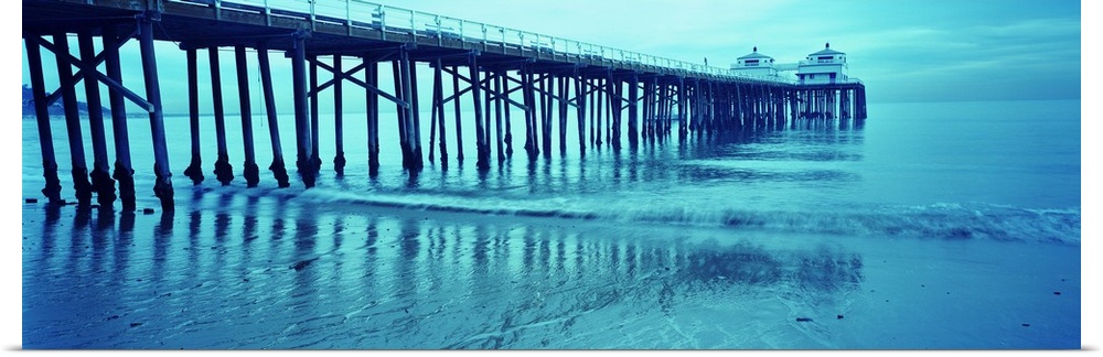 Monotone photograph of a long pier reaching out into the Pacific Ocean at dusk with shallow waves receding as the tide cha...