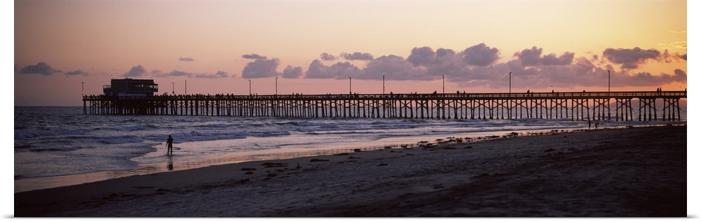 A beach board walk extends out into the ocean in this landscape photograph of the sun setting on a sandy beach.