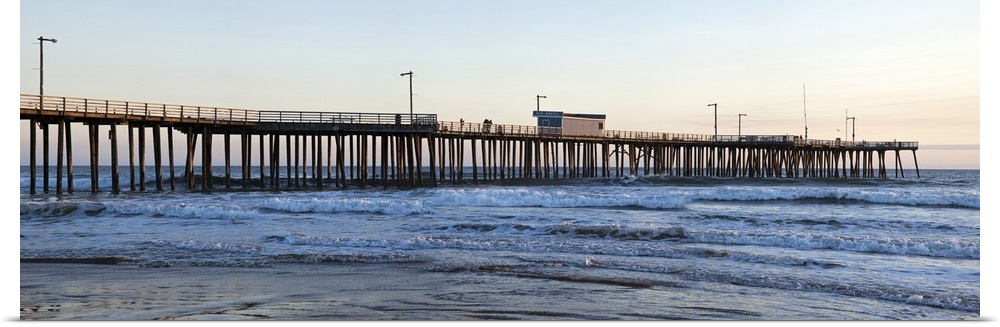 Large landscape photograph of Pismo Beach Pier extending into the waters along Pismo Beach, in San Luis, California, at dusk.