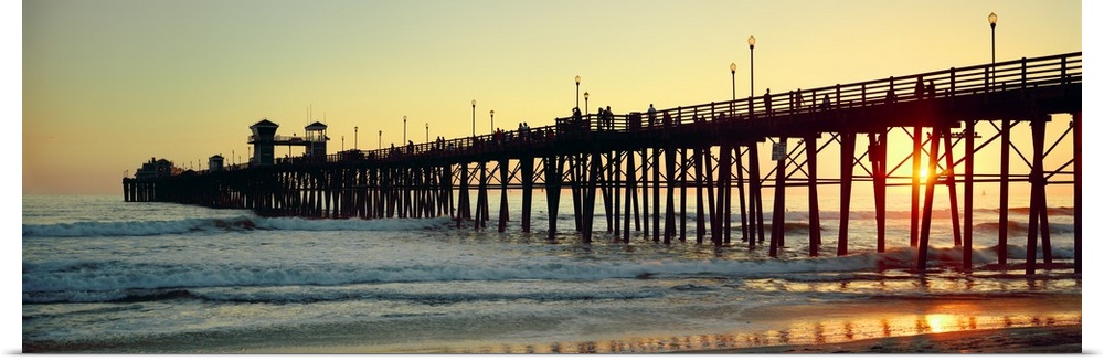 Panoramic wall docor of the silhouette of a pier reaching into the ocean surf at sunset.
