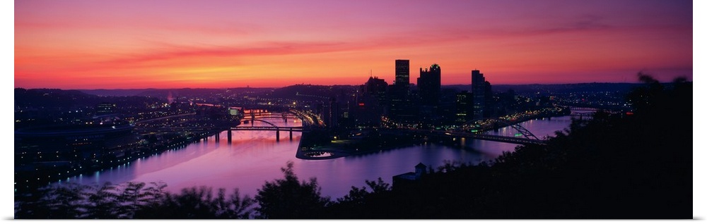 Oversized, landscape photograph of the distant skyline of Pittsburgh, Pennsylvania lit up, beneath a vibrant sunset.