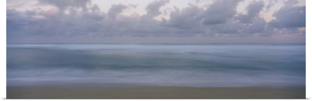 This is a panoramic photograph of a waves gently washing on the sandy shore with a cloudy sky overhead in this seascape.