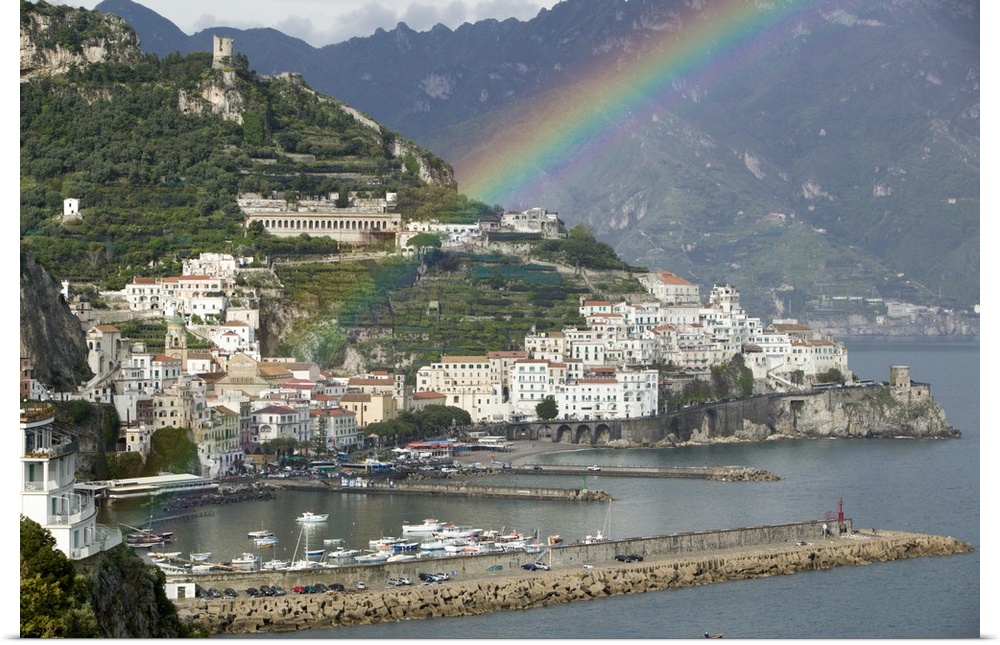 This is a picture of a rainbow over a town off a coast in Italy. Large white buildings line the water that has piers with ...