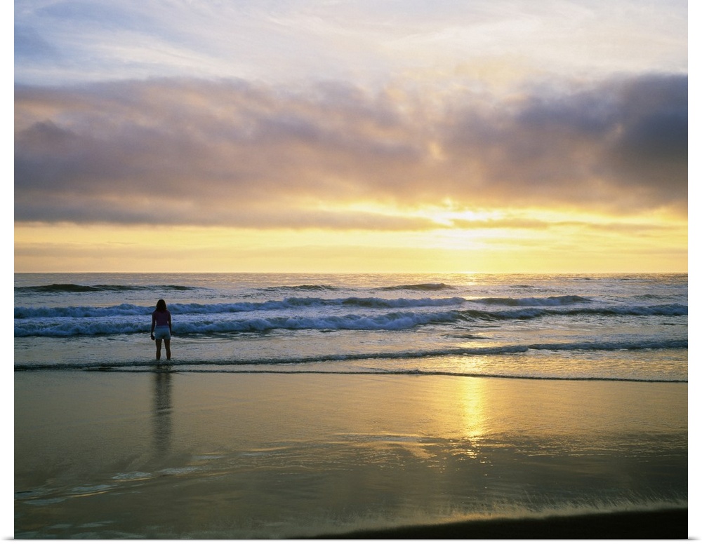 In this landscape photograph a lone figure stands in the waves on the shore watching the sunset over the horizon.