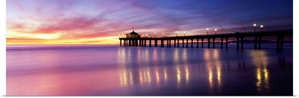 Panoramic photograph of pier extending into ocean at sunset. The lights on the pier are reflected in the ocean and the sky...