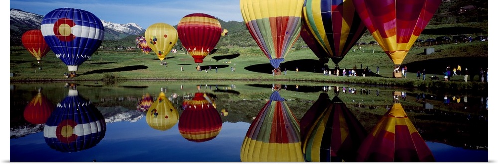 Long photo on canvas of big balloons about to take off in a field that are reflected in the waterfront.