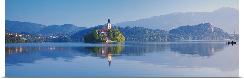 Giant, horizontal photograph of a mountain landscape and buildings reflecting in the waters of Lake Bled, in Slovenia.