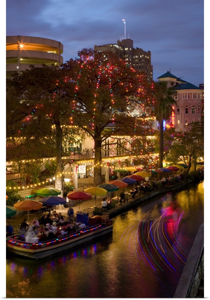 Vertical photo on canvas of people eating at an outdoor restaurant next to a river underneath umbrellas and sparkling lights.