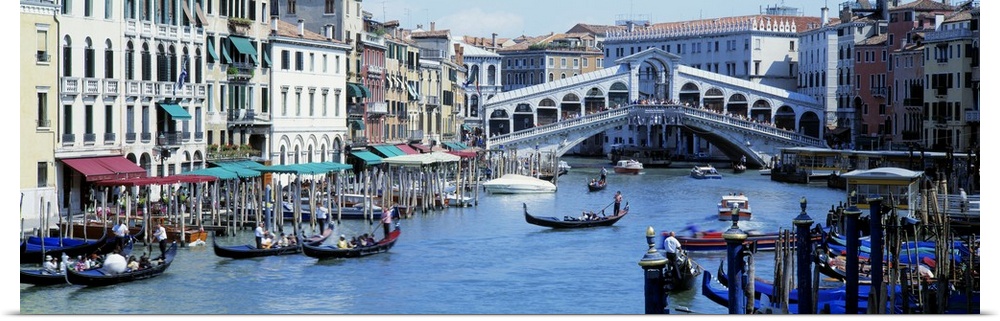 Panoramic photograph of waterway filled with gondolas and lined with shops and buildings, with overpass in distance.