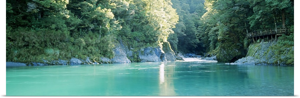 Panoramic image of a river with clear water flowing through a dense forest with big rocks as cliffs on either side.