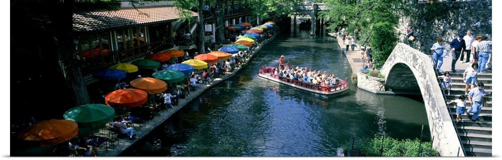 A panoramic picture of a river in Texas that is lined with tables that have colorful umbrellas and a small passenger boat ...
