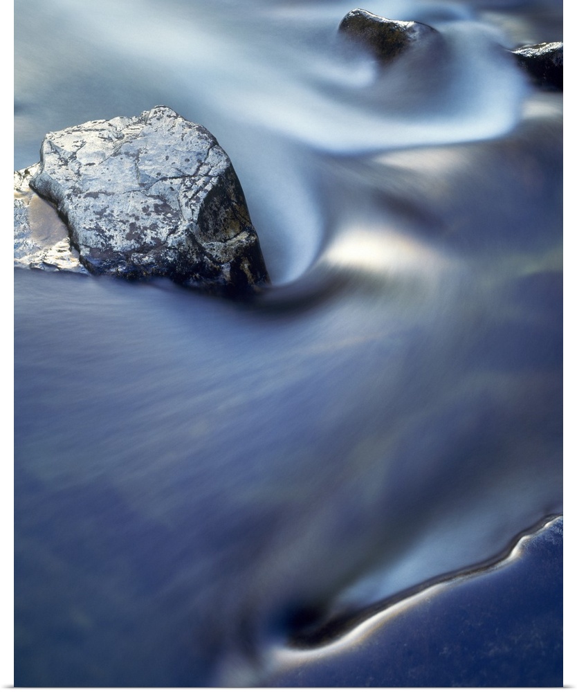 Vertical, close up photograph on a large wall hanging  of blurred water in rapid movement as it swirls around rocks in the...