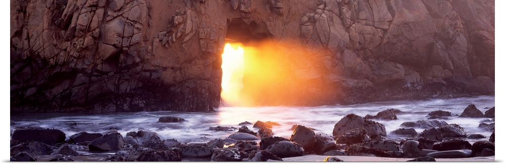 Panoramic photograph of rocky shoreline with sun peering through huge stone arch-like structure.