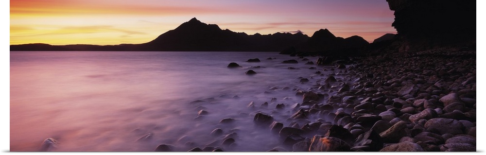 Panoramic photograph of pebble covered shoreline rising from mist with mountain silhouette in the distance at sunset.