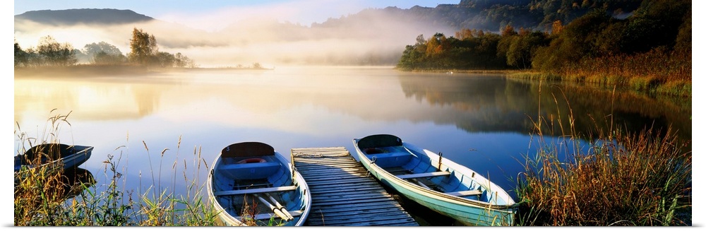 Panoramic photograph showcases rowboats sitting next to a dock on a lake in Grasmere within Cumbria, England.  In the back...