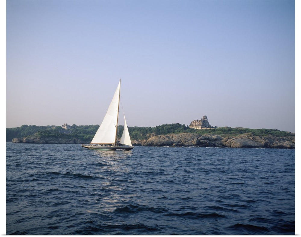 A single sail boat drifts in the ocean. A large home is pictured on land behind the boat and rocky coast.