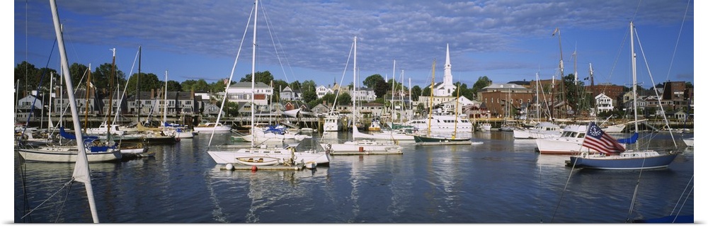 This is a panoramic photograph of a harbor in New England filled with sail boats and a small village in the background.