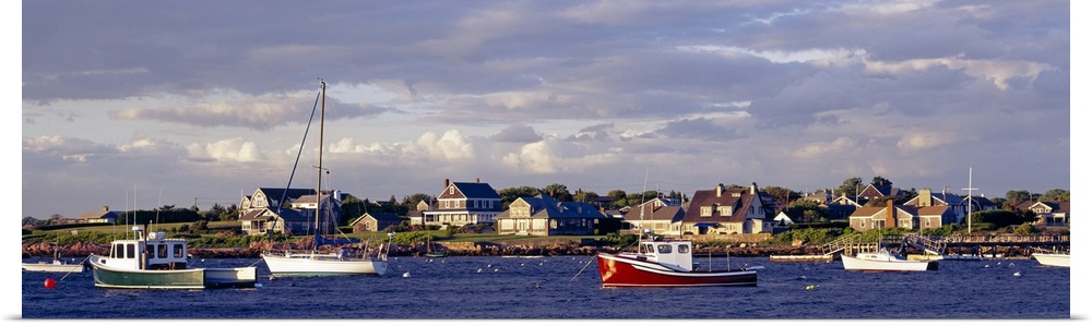 Large landscape photograph of many boast in Sakonnet Harbor, houses line the landscape, beneath a mostly cloudy sky in Rho...