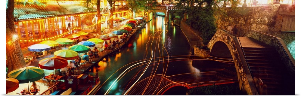 Nighttime panoramic photo of the Riverwalk in San Antonio as boats travel through the canals and people eat under colorful...