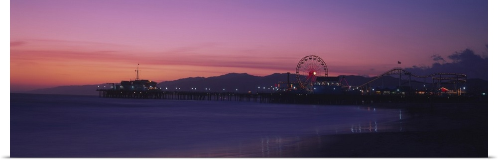 A panoramic photograph of a board walk, Ferris wheel, and rollercoaster on a board walk during a pastel sunset.