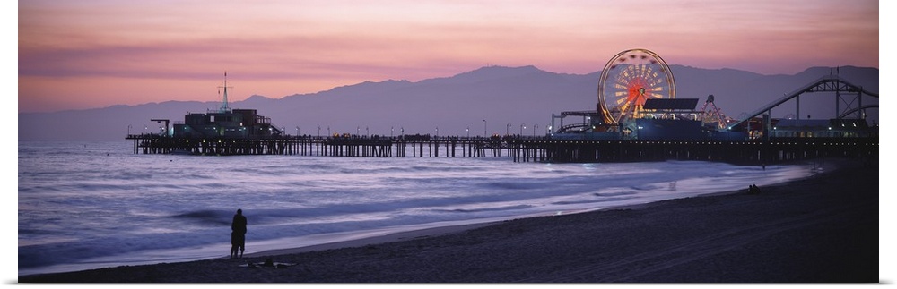 A panoramic shot taken of the Santa Monica pier during sun down with a couple standing on the beach at the waters edge.